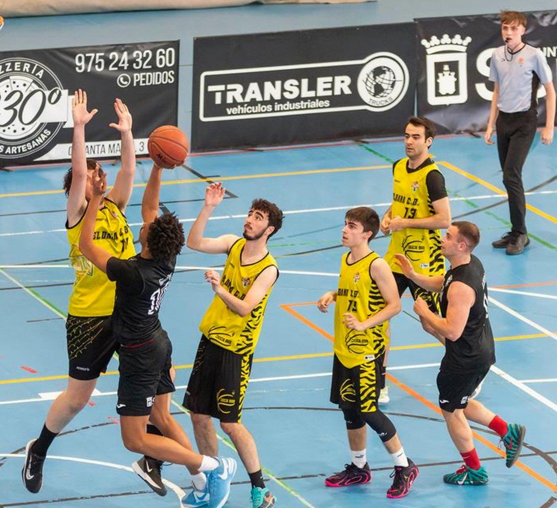 Basketball game in progress: players in yellow and black jerseys reach for the ball on an indoor court with banners in the background