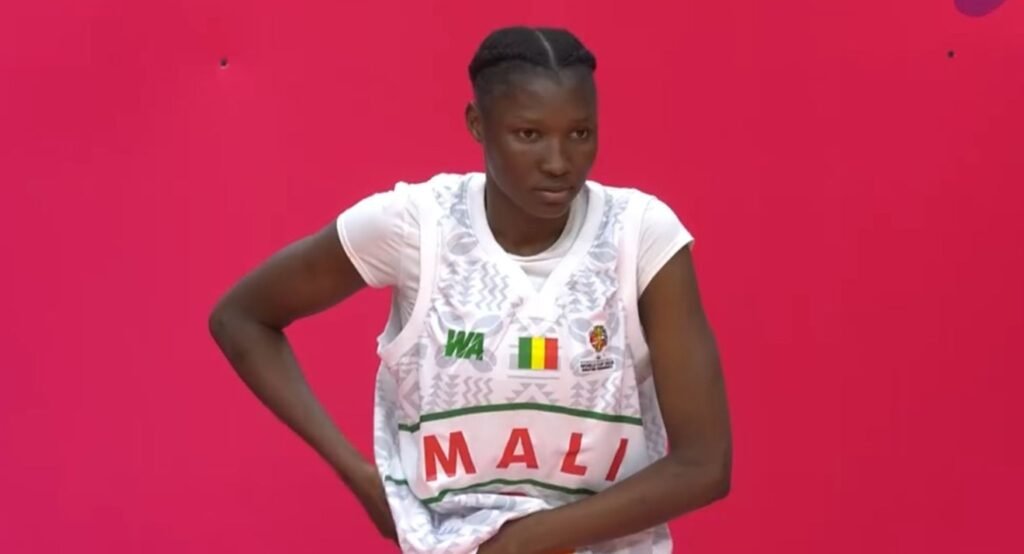 Female athlete wearing a white Mali jersey with the Malian flag, standing with hands on her hips against a bright pink background.