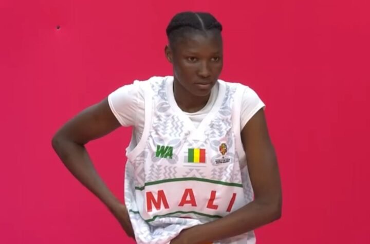 Female athlete wearing a white Mali jersey with the Malian flag, standing with hands on her hips against a bright pink background.