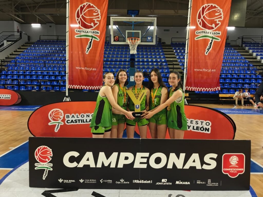 Girls basketball team posing with a trophy after a win, on a gym floor with banners reading Castilla y León and CAMPEONAS in the foreground.