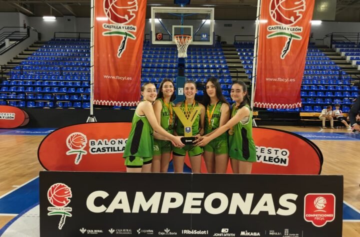 Girls basketball team posing with a trophy after a win, on a gym floor with banners reading Castilla y León and CAMPEONAS in the foreground.