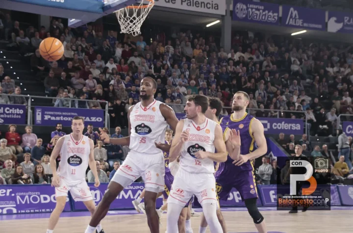 Basketball players contesting under the hoop as the ball flies toward the basket; crowded stands in the background.