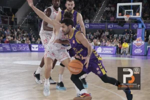 Basketball player in purple drives with the ball past a defender in white on a crowded court.