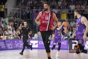 Basketball player in a red jersey (#13) running down the court as a game unfolds with fans watching from the stands behind him.