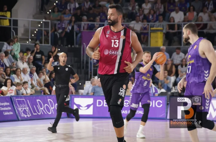 Basketball player in a red jersey (#13) running down the court as a game unfolds with fans watching from the stands behind him.
