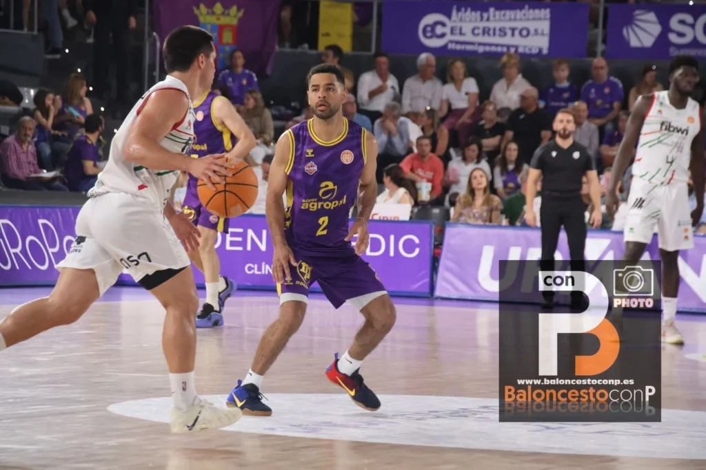 Basketball game: a purple-yellow team player guards a ball handler while a white-uniform defender approaches on a glossy court, with spectators in the stands behind.