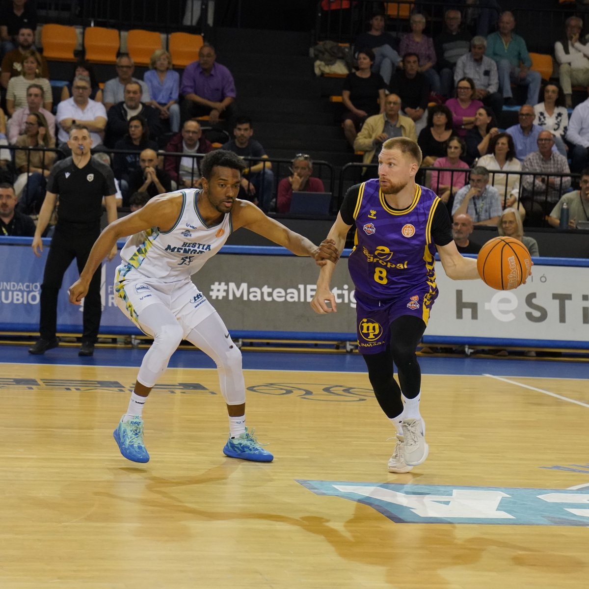 Purple-jerseyed guard (number 8) dribbles past a white-jersey defender as fans watch from the stands.