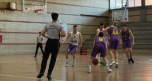Indoor basketball game: players in purple and white uniforms contest the ball near the key while a referee watches from the foreground.