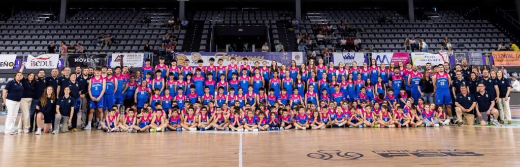 Large group of children and adults in colorful basketball uniforms posing for a team photo on an indoor court.