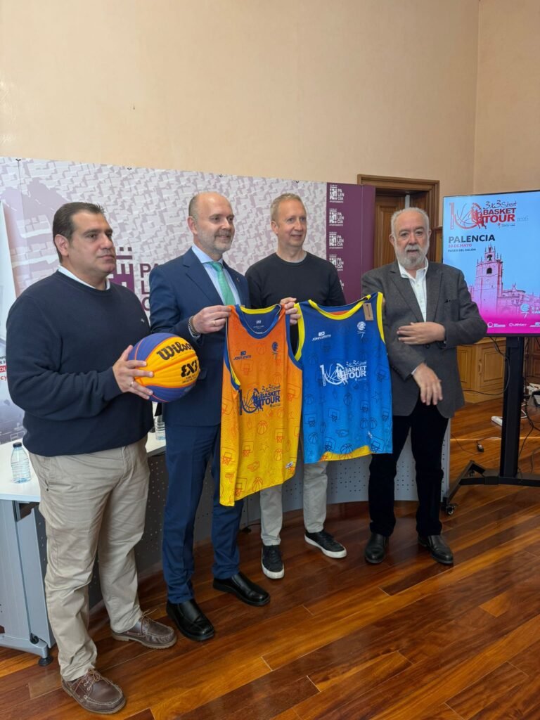 Four men posing with orange and blue basketball jerseys at a press event, holding the jerseys up for display in front of a banner and screen.