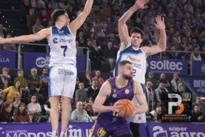 A basketball player in purple drives with the ball as two defenders in white jerseys leap to block him in a packed arena crowd behind them.
