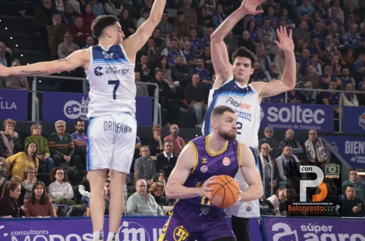 A basketball player in purple drives with the ball as two defenders in white jerseys leap to block him in a packed arena crowd behind them.