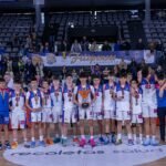 Youth basketball team posing with medals on the court, audience and banners in the stands behind them, trophy in center.