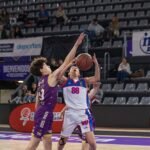 Two male basketball players reach for the ball while a teammate in purple guards near them in an indoor court with empty stands in the background.