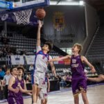 Basketball players jump near the hoop as a white-jersey player (#2) goes for a layup and a purple-jersey defender (#10) challenges him.