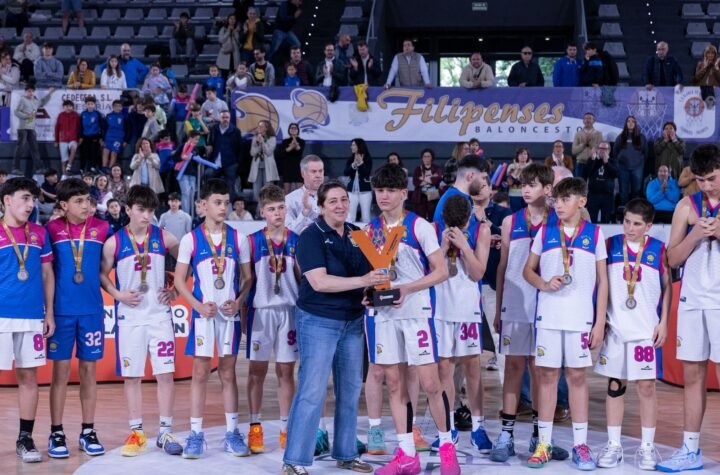 Young basketball players in blue and white uniforms stand in a row on the court as a woman hands a player a trophy, with spectators watching from the stands behind them.
