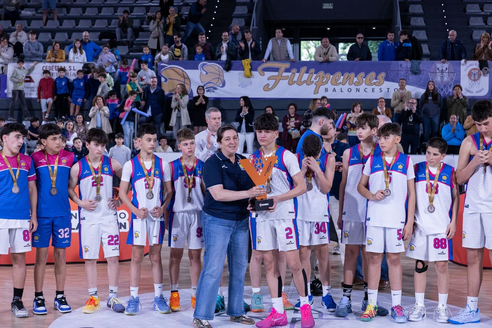 Young basketball players in blue and white uniforms stand in a row on the court as a woman hands a player a trophy, with spectators watching from the stands behind them.