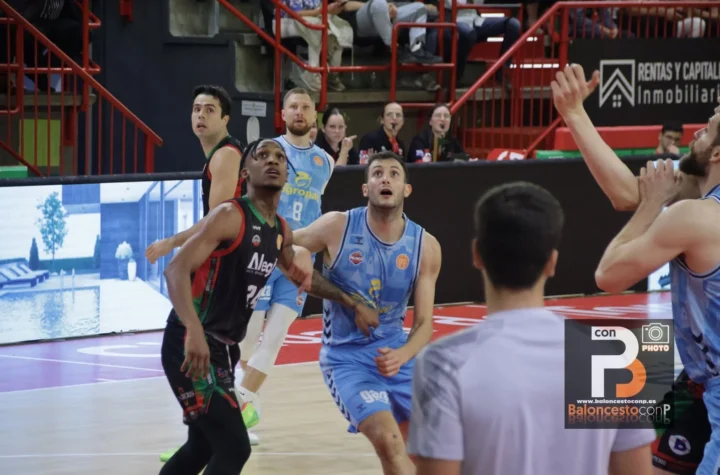 Basketball players in blue and dark jerseys look upward as they chase the ball near the basket, with red arena rails and spectators in the background.