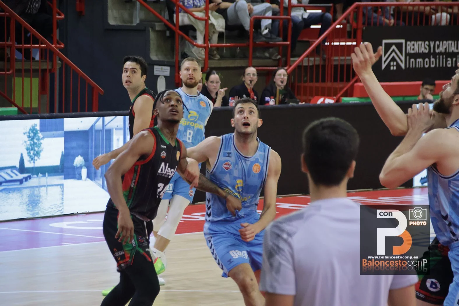 Basketball players in blue and dark jerseys look upward as they chase the ball near the basket, with red arena rails and spectators in the background.