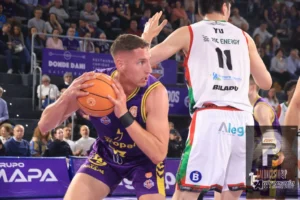 Basketball player in purple drives with the ball while being guarded by a defender in white jersey; crowd watching from stands in the background.
