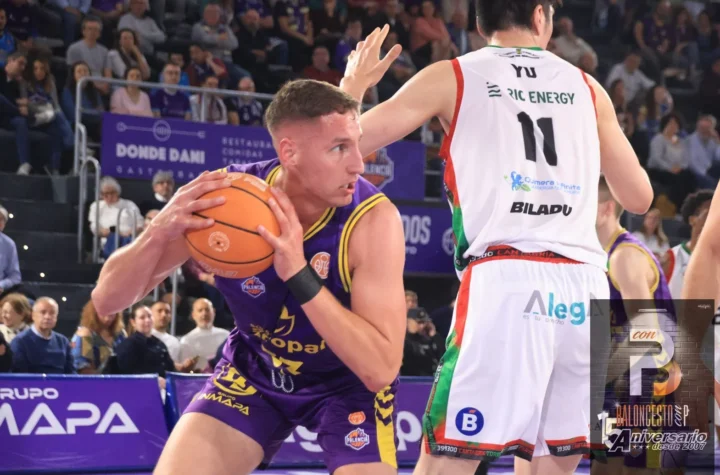 Basketball player in purple drives with the ball while being guarded by a defender in white jersey; crowd watching from stands in the background.