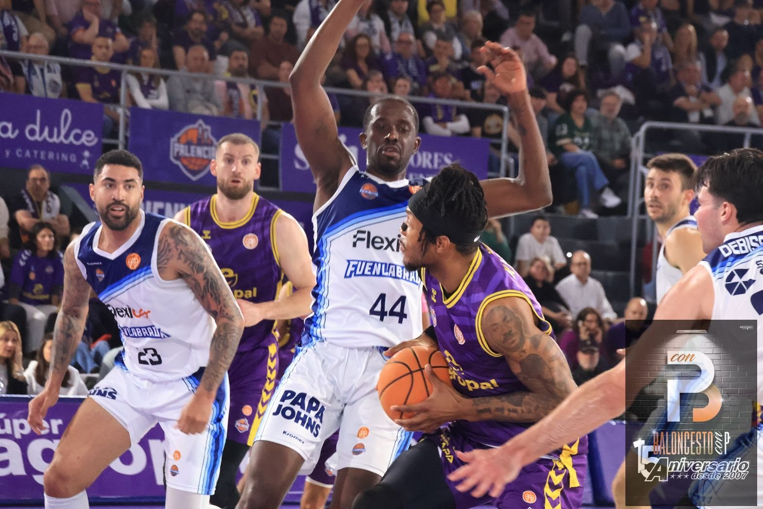 Basketball game in progress: a purple-uniform player drives with the ball while white-uniform opponents converge, crowd in stands behind.