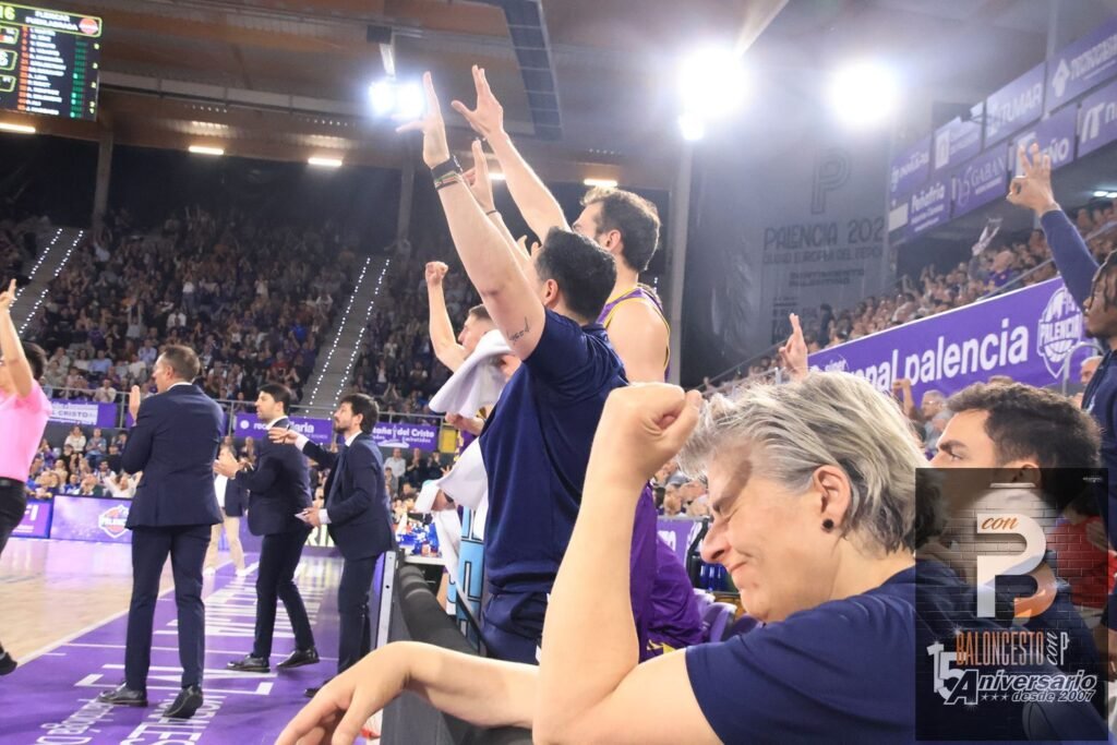 Basketball players and coaches celebrate on the sideline with arms raised as fans cheer in a purple-clad arena.