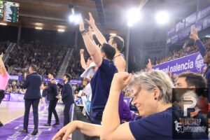 Basketball players and coaches celebrate on the sideline with arms raised as fans cheer in a purple-clad arena.