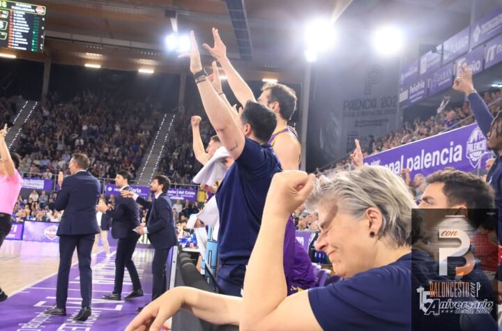 Basketball players and coaches celebrate on the sideline with arms raised as fans cheer in a purple-clad arena.