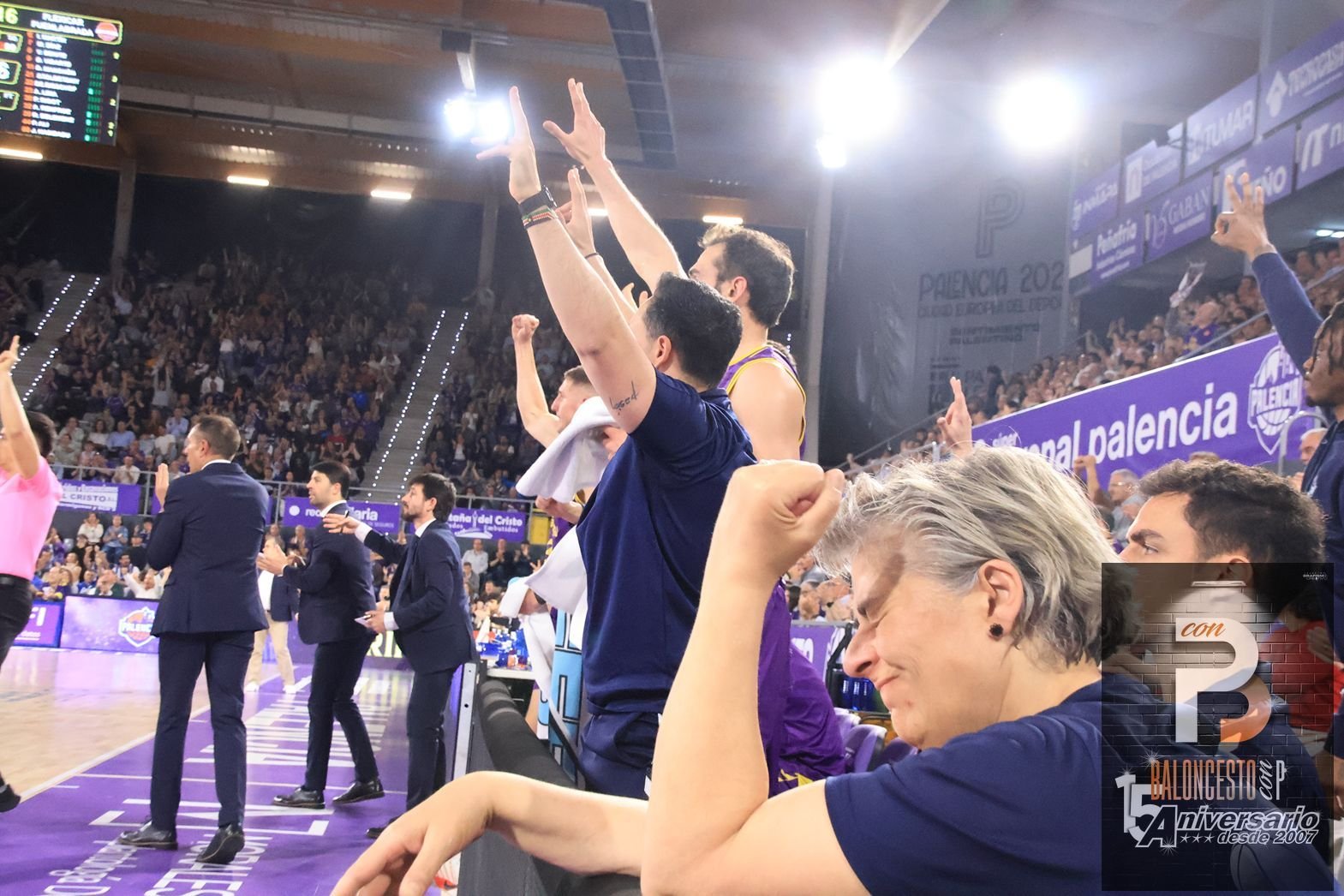 Basketball players and coaches celebrate on the sideline with arms raised as fans cheer in a purple-clad arena.