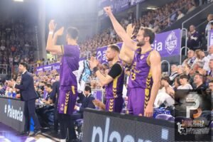 Basketball players in purple uniforms stand and cheer on the sideline as the crowd applauds in the arena.