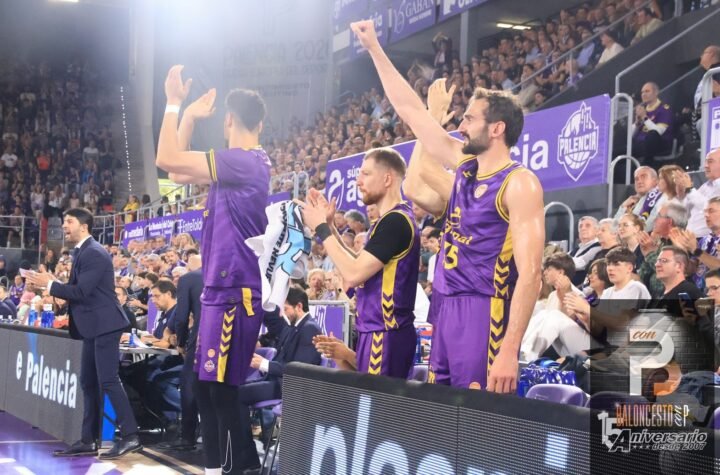 Basketball players in purple uniforms stand and cheer on the sideline as the crowd applauds in the arena.