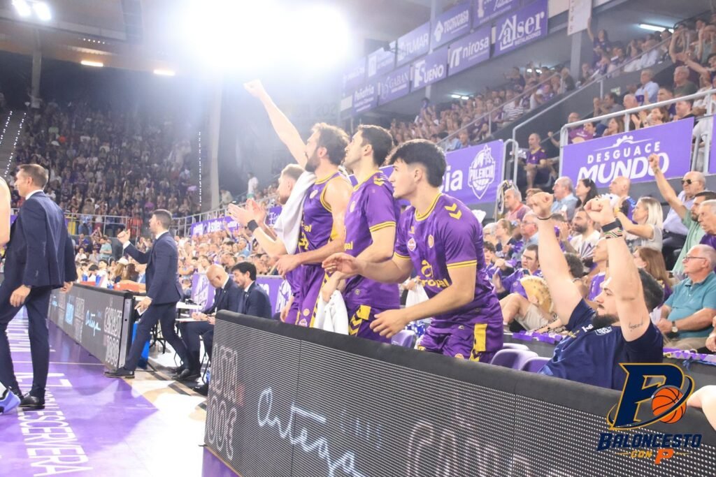 Basketball players in purple uniforms standing along the sideline, cheering with fans in the stands behind them