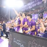 Basketball players in purple uniforms standing along the sideline, cheering with fans in the stands behind them