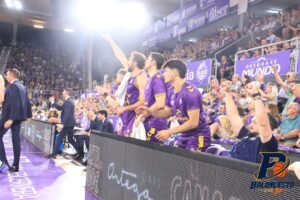 Basketball players in purple uniforms standing along the sideline, cheering with fans in the stands behind them
