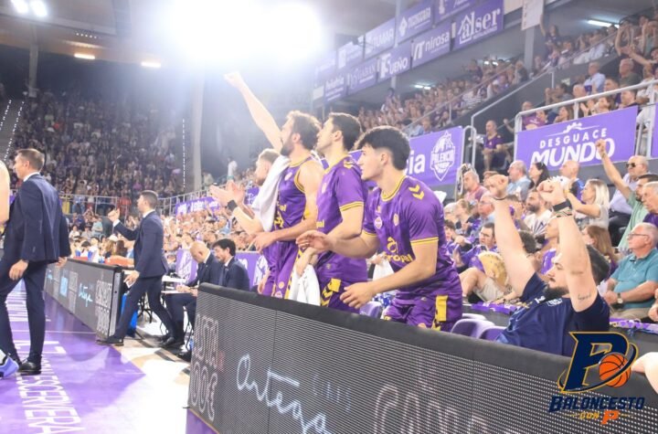 Basketball players in purple uniforms standing along the sideline, cheering with fans in the stands behind them