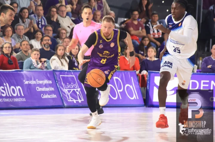 Basketball player in purple and yellow jersey dribbles fast toward the basket while a defender in white watches in a crowded arena.