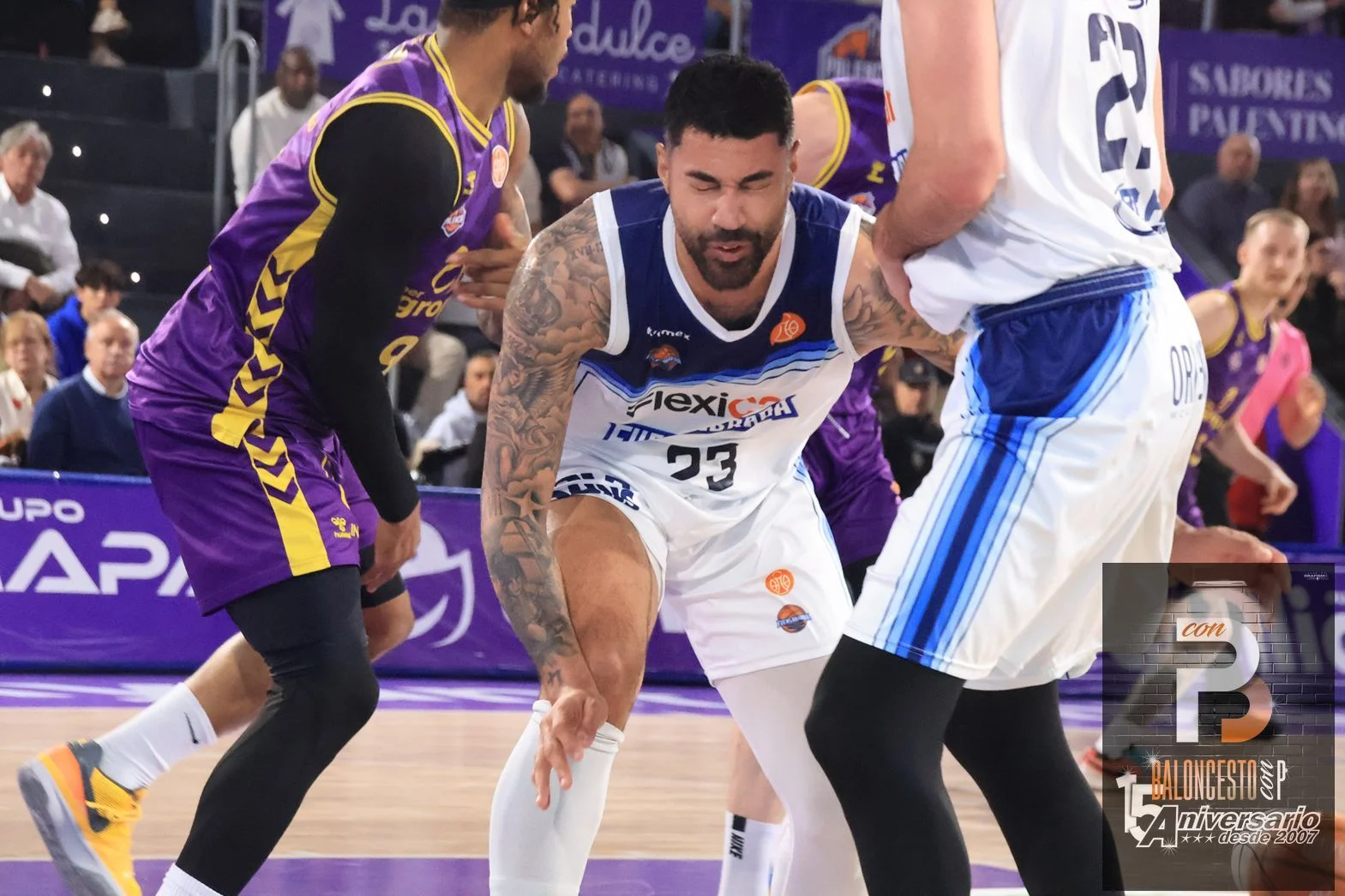 Basketball players in purple and white uniforms contest a ball near the court; central player guards the ball while teammates surround him.