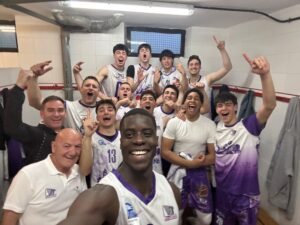 Group of basketball players in a locker room posing for a celebratory selfie, fists raised and smiles on their faces in white and purple uniforms.