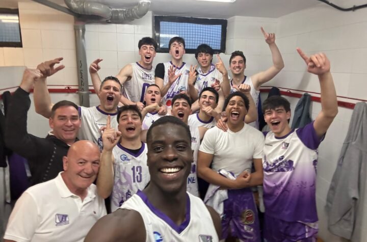 Group of basketball players in a locker room posing for a celebratory selfie, fists raised and smiles on their faces in white and purple uniforms.