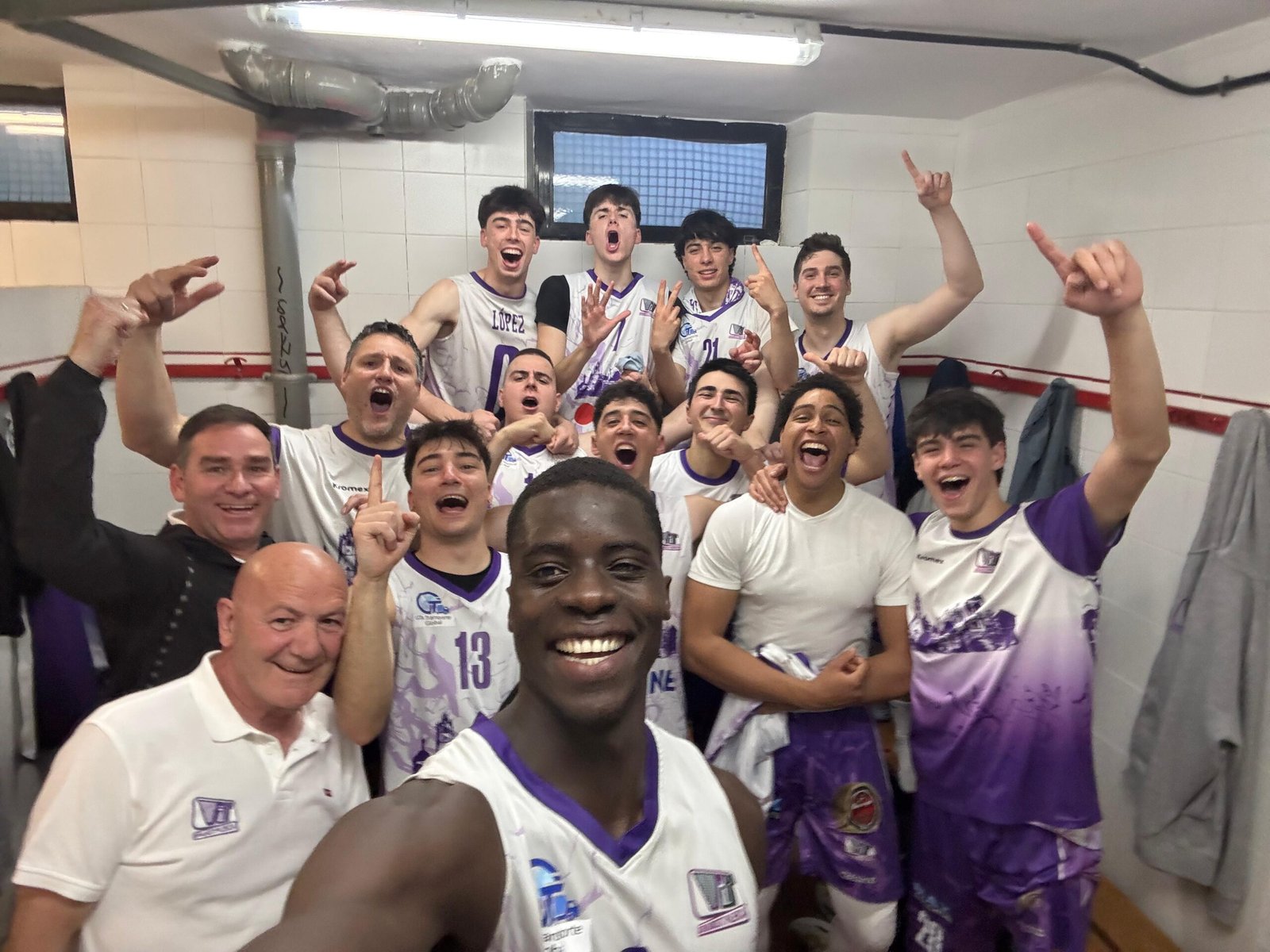 Group of basketball players in a locker room posing for a celebratory selfie, fists raised and smiles on their faces in white and purple uniforms.