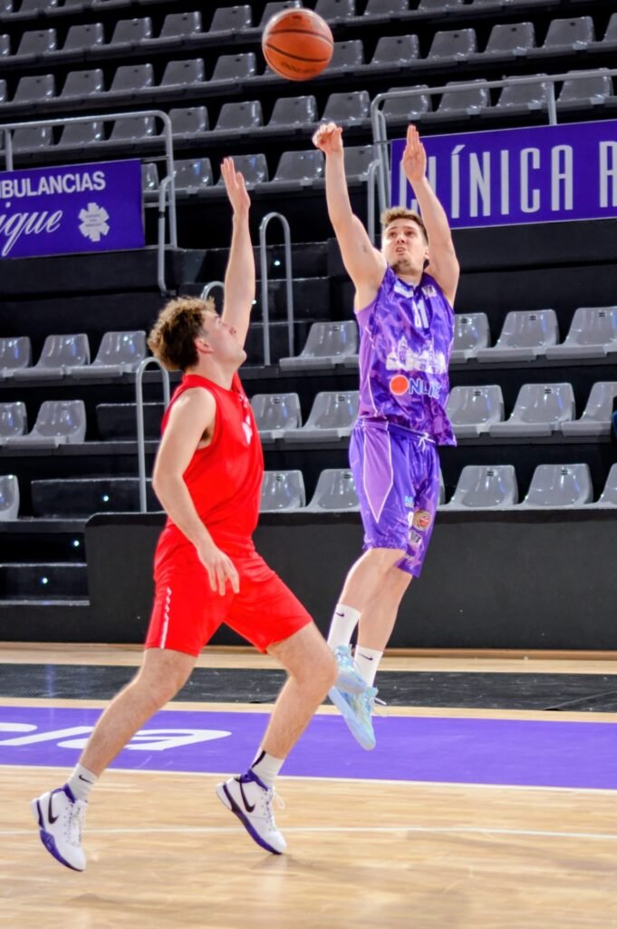 Two male basketball players in mid-air contesting the ball during a game in an indoor arena; one player in red jersey reaches up while the other in purple jumps for the ball, with empty spectator seats in the background.