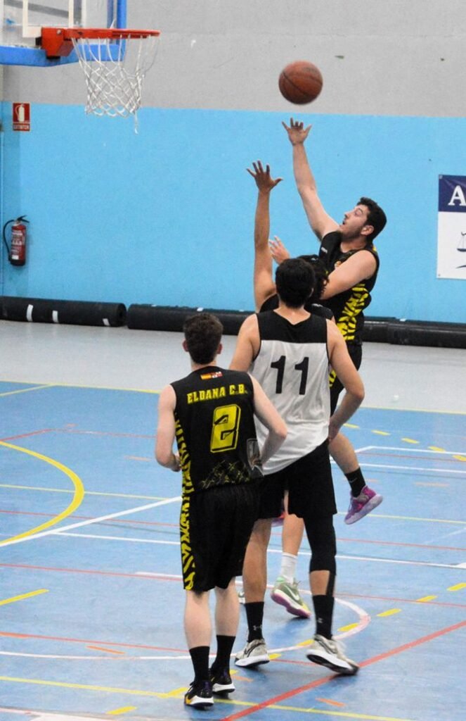 Basketball game in progress: a player in a black-yellow jersey soars for a shot near the hoop while two defenders reach up nearby.