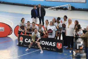 Female basketball players celebrate a victory on a blue indoor court, wearing white jerseys with medals and a trophy, standing behind a sponsor banner with coaches and supporters nearby.