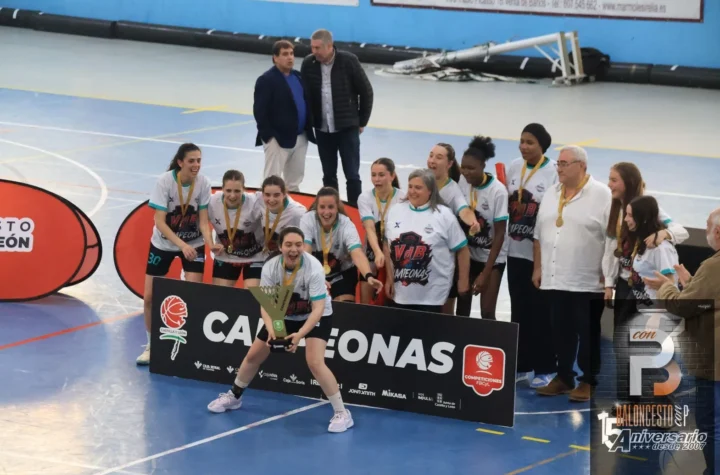 Female basketball players celebrate a victory on a blue indoor court, wearing white jerseys with medals and a trophy, standing behind a sponsor banner with coaches and supporters nearby.