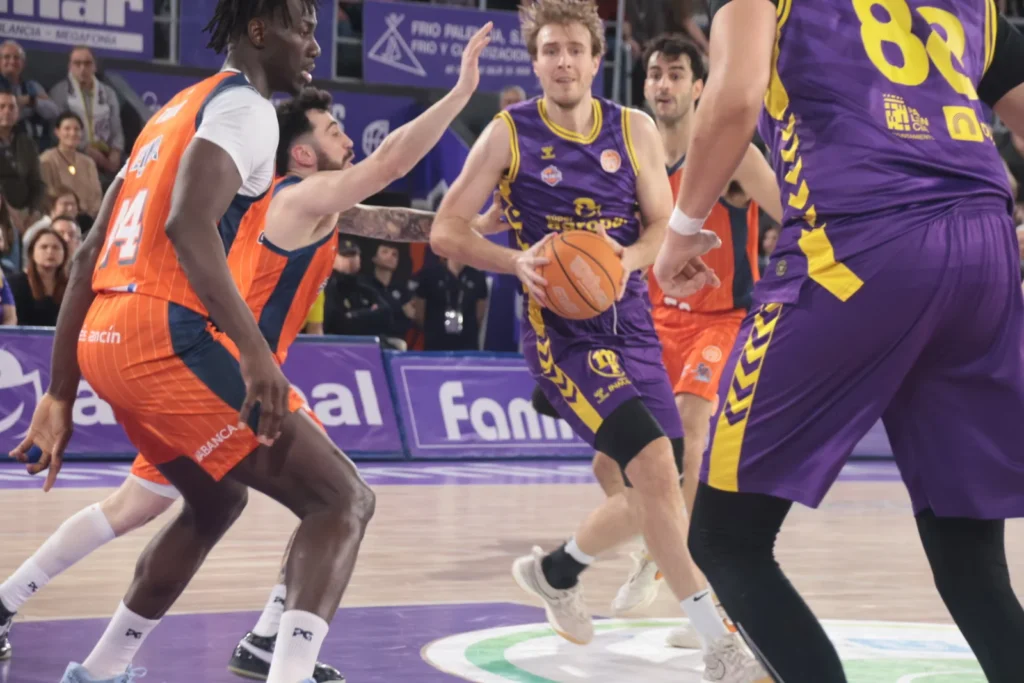 Basketball player in purple holds the ball while an orange-clad defender reaches in, during a fast-paced move on the court.