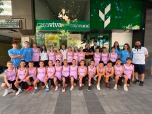 Group of young basketball players in pink uniforms posing with coaches outside a bank storefront.