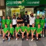 Group of young female athletes in green shirts posing with coaches outside a Caja Rural storefront.
