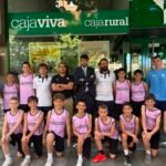 Group of young basketball players in pink jerseys posing with coaches outside a green CajaViva storefront.