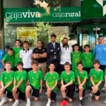 Group of young soccer players in green shirts pose with coaches outside a bank storefront with a green sign reading 'caja viva' and 'cajarural'.